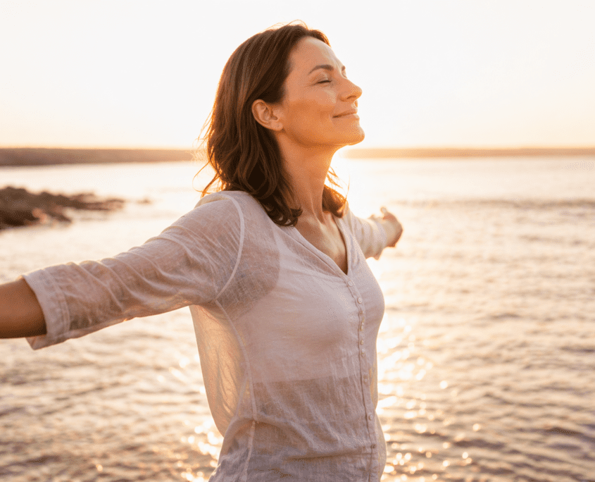 Woman stands with arms outstretched at sunset over a calm body of water, eyes closed in contentment.