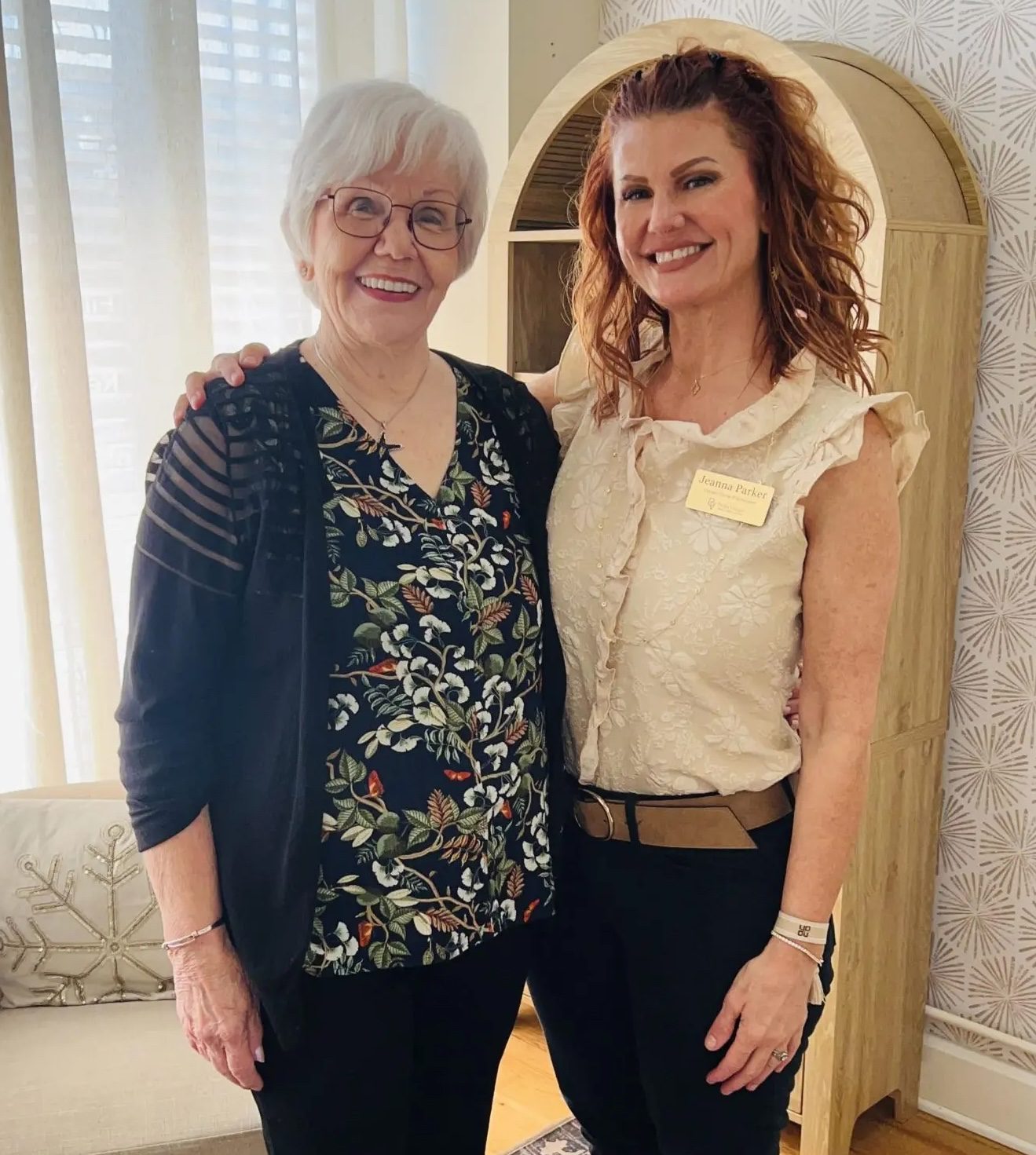 Two women posing indoors: an older woman with white hair and glasses in a floral top, and a younger woman with auburn hair in a cream lace blouse wearing a name badge.
